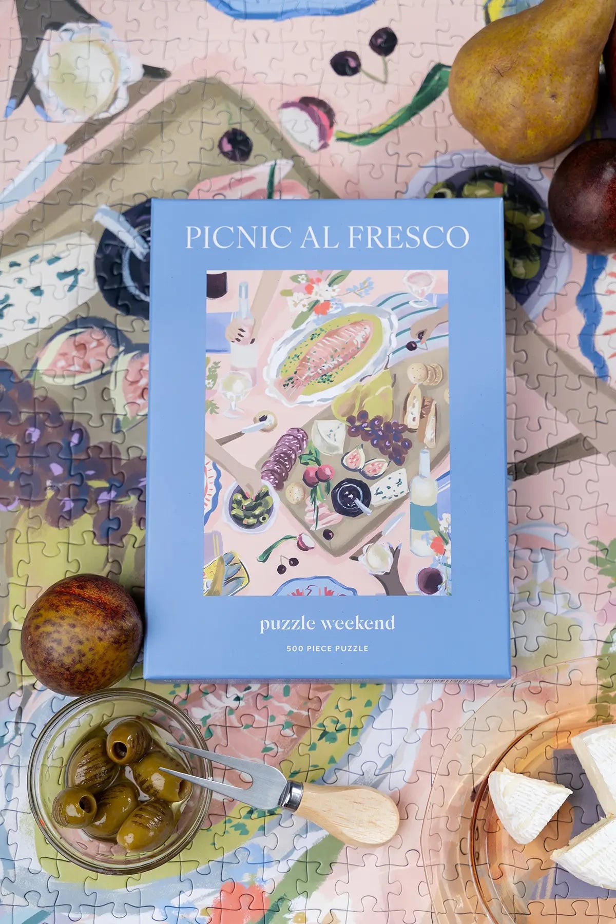 Jigsaw puzzle box titled 'Picnic al Fresco' on a partially completed puzzle with fruits and vegetables.